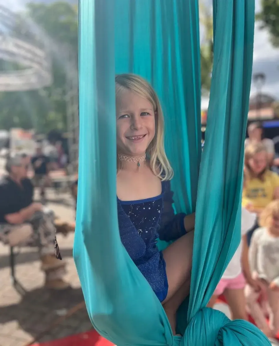 Niña sonriendo en una hamaca de tela turquesa en un evento al aire libre.