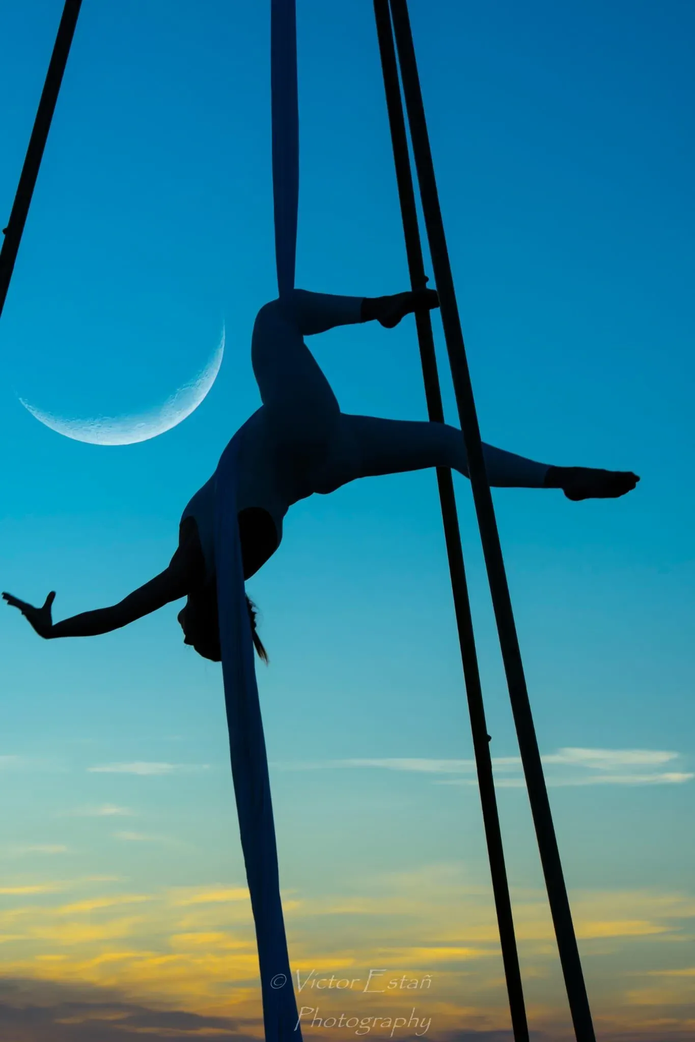 Silueta de un artista aéreo suspendido de cuerdas contra un cielo de puesta de sol con una luna creciente.