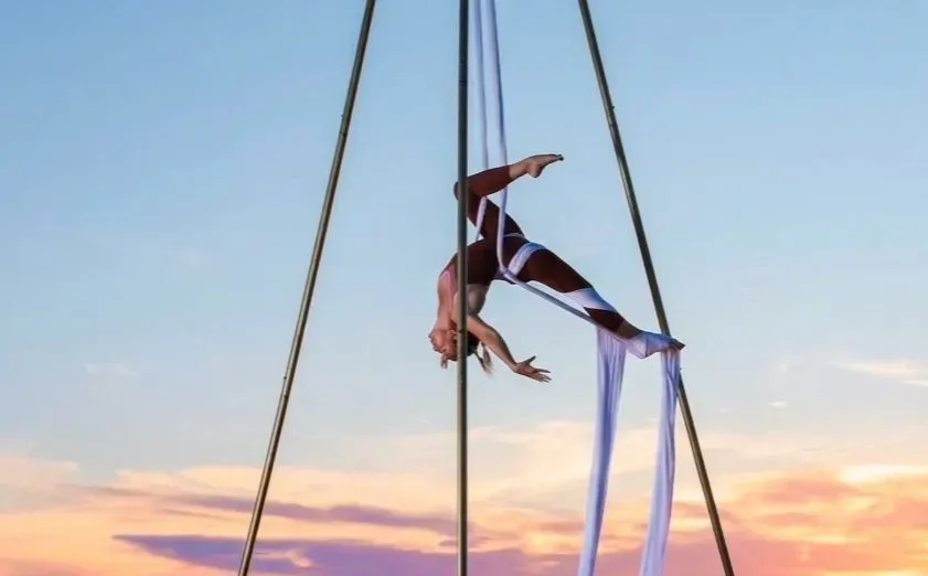 Artista aérea suspendida boca abajo entre telas bajo un cielo de atardecer en tonos pastel.