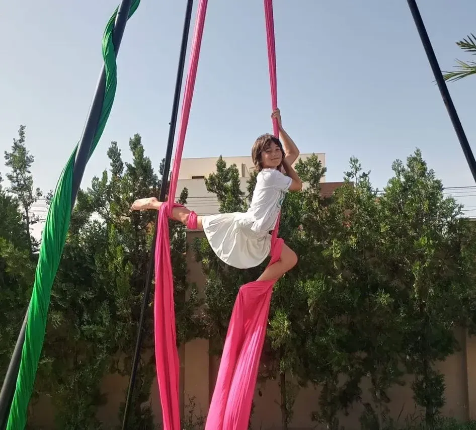 Una persona sobre una tela aérea rosa al aire libre, sonriendo y posando con un fondo de árboles y un edificio.