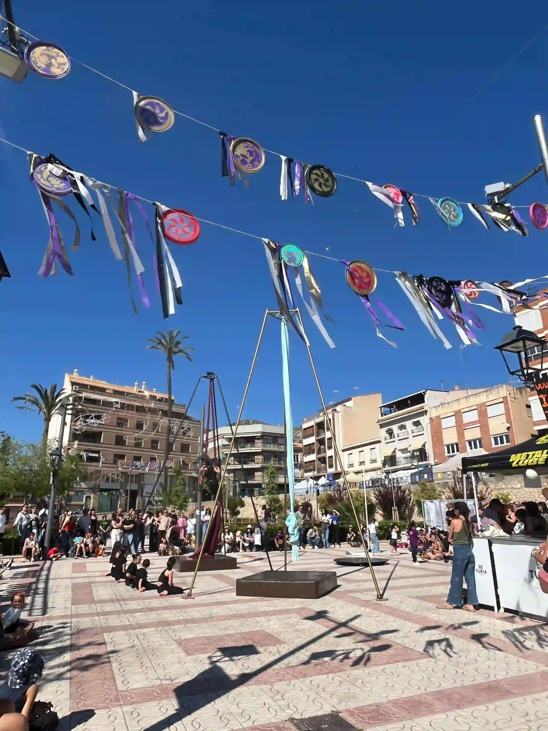 Plaza decorada con banderas ondeando, gente reunida y una estructura de arco blanco bajo un cielo azul brillante.