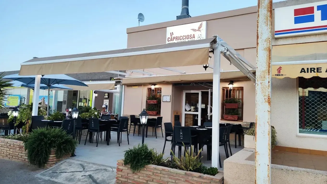 Cafetería al aire libre con toldo beige, mesas y sillas negras y un pequeño patio frente a un local comercial.