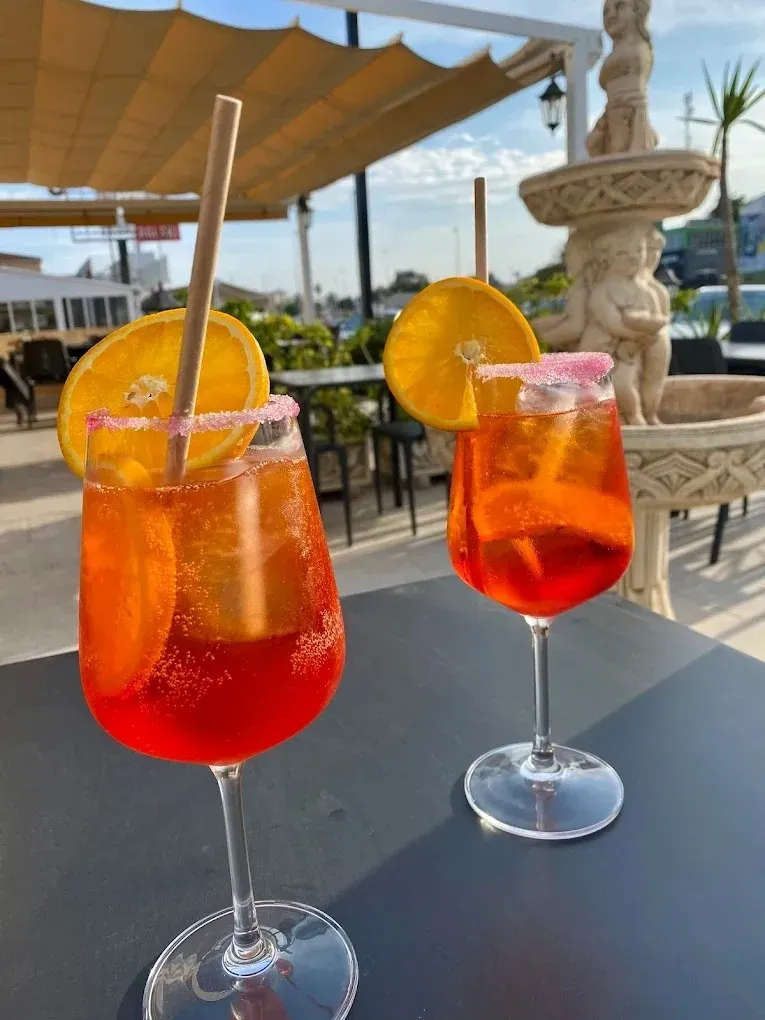 Dos cócteles de naranja en copas con tallo sobre una mesa de patio junto a la playa.