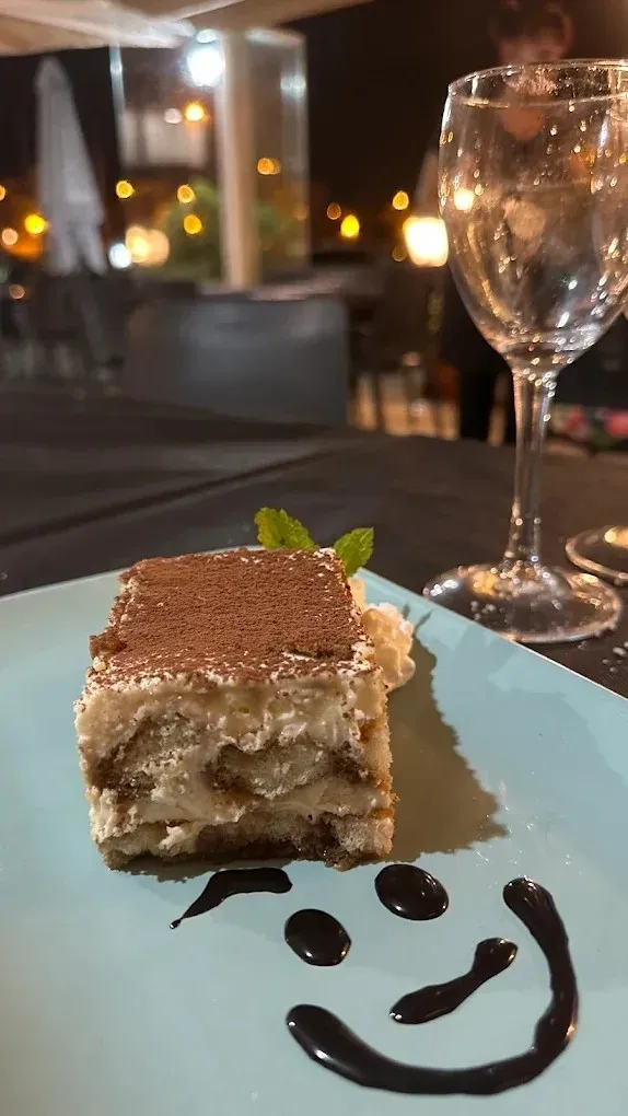 Postre en un plato azul con una carita sonriente de chocolate, junto a un vaso en un ambiente de restaurante con luz tenue.