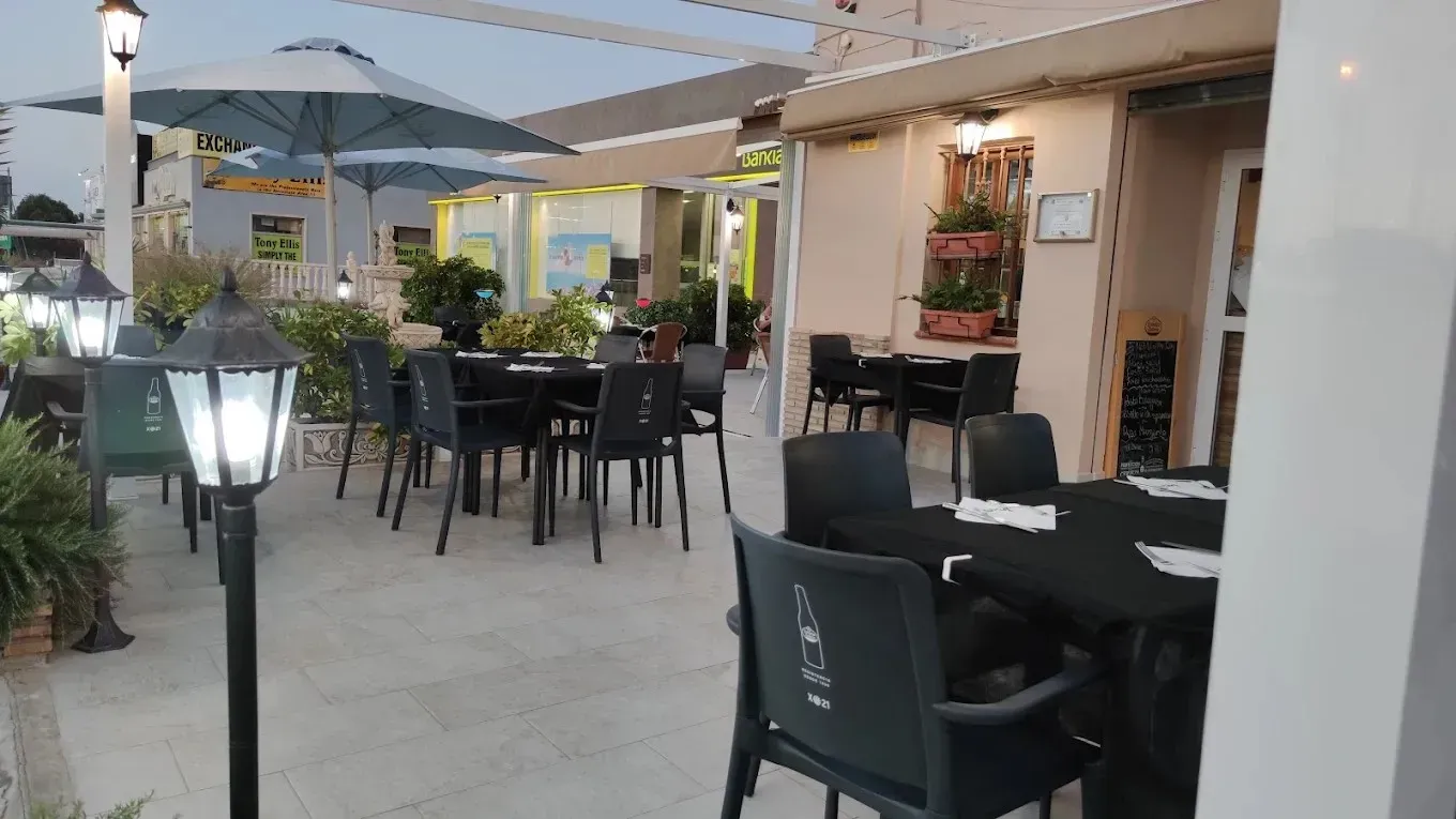 Zona de comedor al aire libre en una cafetería, con sillas negras, mesas y grandes sombrillas.