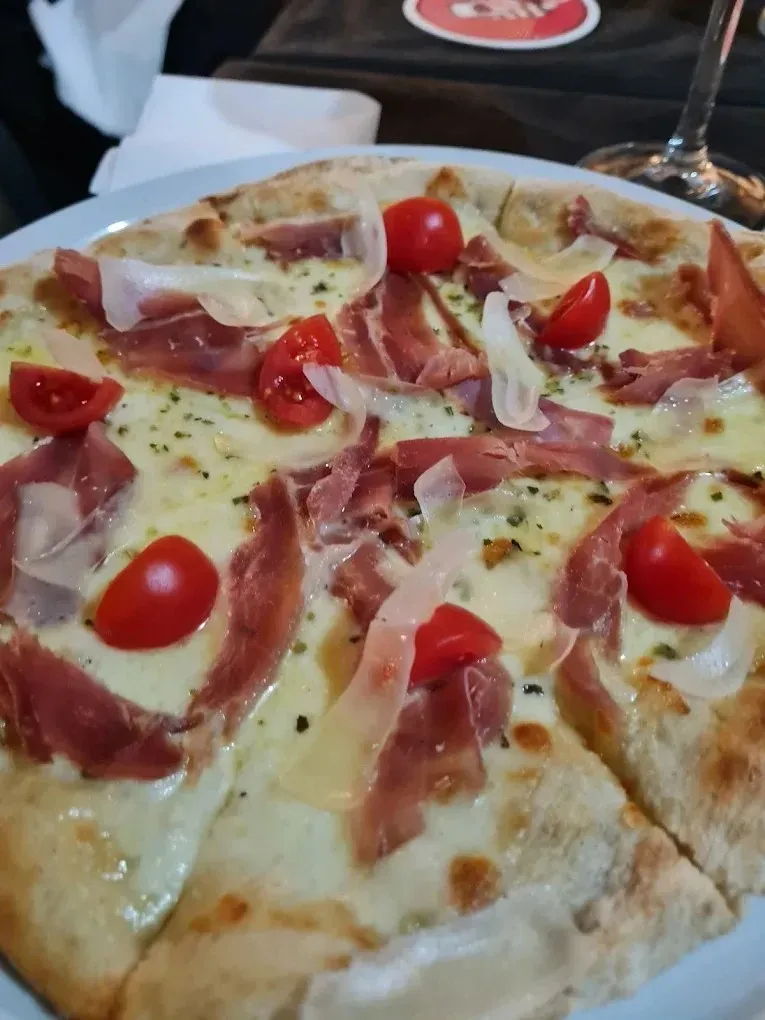 Pizza con tomates cherry, jamón serrano, queso fundido y hierbas aromáticas sobre una mesa.