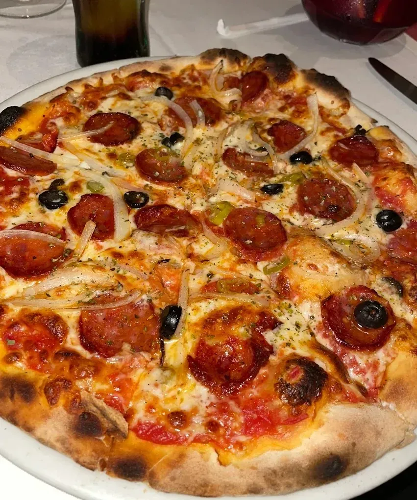 Pizza de pepperoni, aceitunas y champiñones en un plato, recién salida del horno, con una bebida al fondo.
