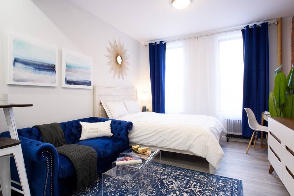 Modern studio apartment with a white bed, blue armchair, clear coffee table, large windows with blue curtains, and abstract wall art. The apartment has a washer and dryer and is located in the Upper East Side.