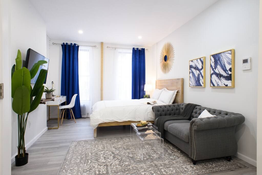 Contemporary studio apartment with a white bed, gray sofa, clear coffee table, potted plant, wall-mounted TV, large windows with blue curtains, and abstract wall art. The apartment has a washer and dryer and is located in the Upper East Side.