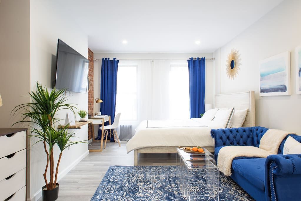 Spacious studio apartment with a white bed, blue armchair, clear coffee table, potted plant, wall-mounted TV, large windows with blue curtains, and abstract wall art. The apartment has a washer and dryer and is located in the Upper East Side.