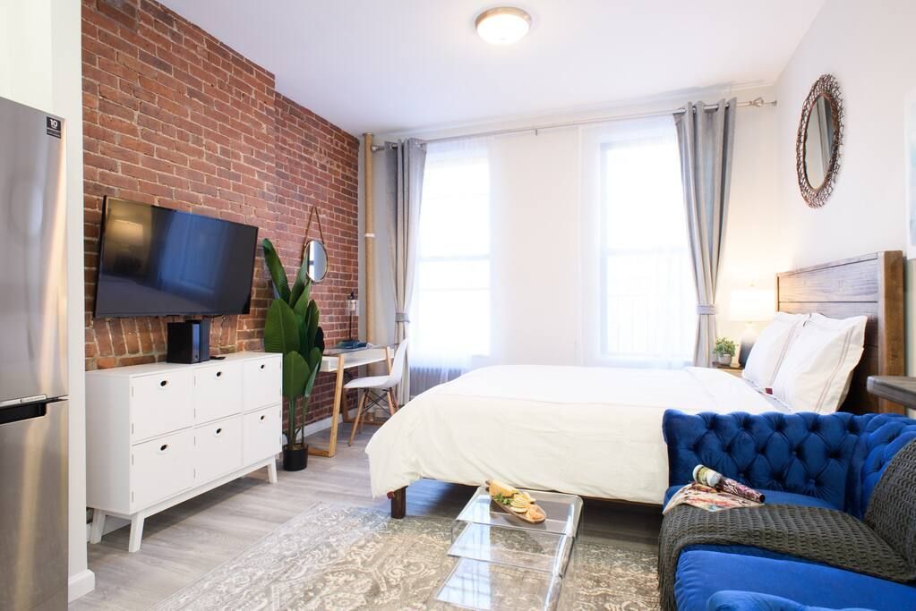 Modern studio apartment with a white bed, blue armchair, clear coffee table, potted plant, wall-mounted TV, large windows with white curtains, and exposed brick walls. The apartment has a washer and dryer and is located in the Upper East Side.