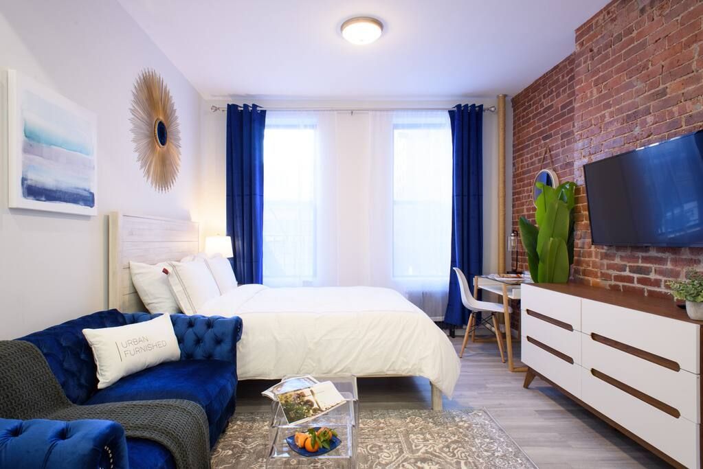 Bright studio apartment with a white bed, blue armchair, clear coffee table, potted plant, wall-mounted TV, large windows with blue curtains, and exposed brick walls. The apartment has a washer and dryer and is located in the Upper East Side.