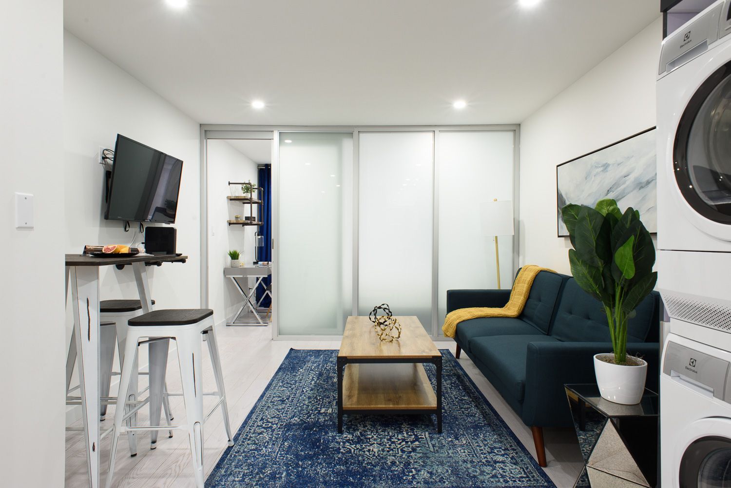 Contemporary one-bedroom apartment with a yellow and gray sofa, blue patterned rug, TV mounted on the wall, breakfast bar with stools, and a small desk. The apartment has a washer and dryer and an in-unit elevator.