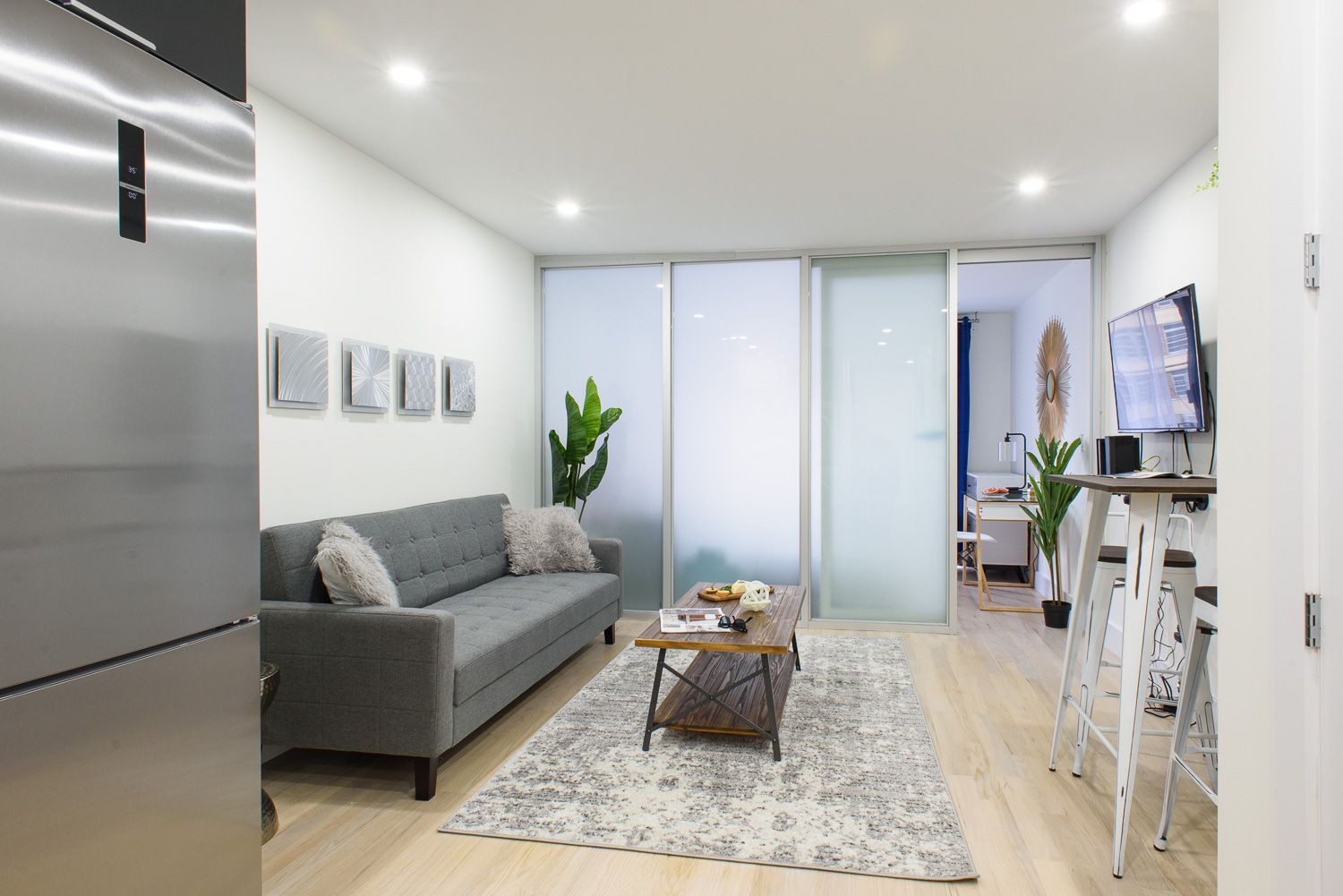 Modern one-bedroom apartment with a gray sofa, glass coffee table, TV mounted on the wall, breakfast bar with stools, and large frosted glass doors. The apartment has a washer and dryer.