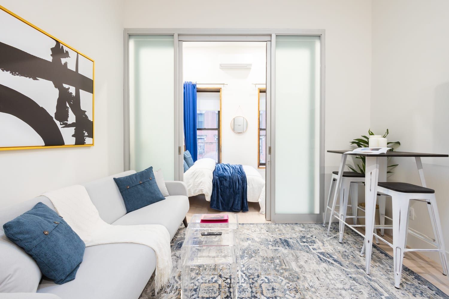 Modern one-bedroom apartment with a white sofa, blue throw pillows, clear coffee table, and a blue patterned rug. The bedroom has a white bed and large frosted glass doors. The apartment has a washer and dryer.
