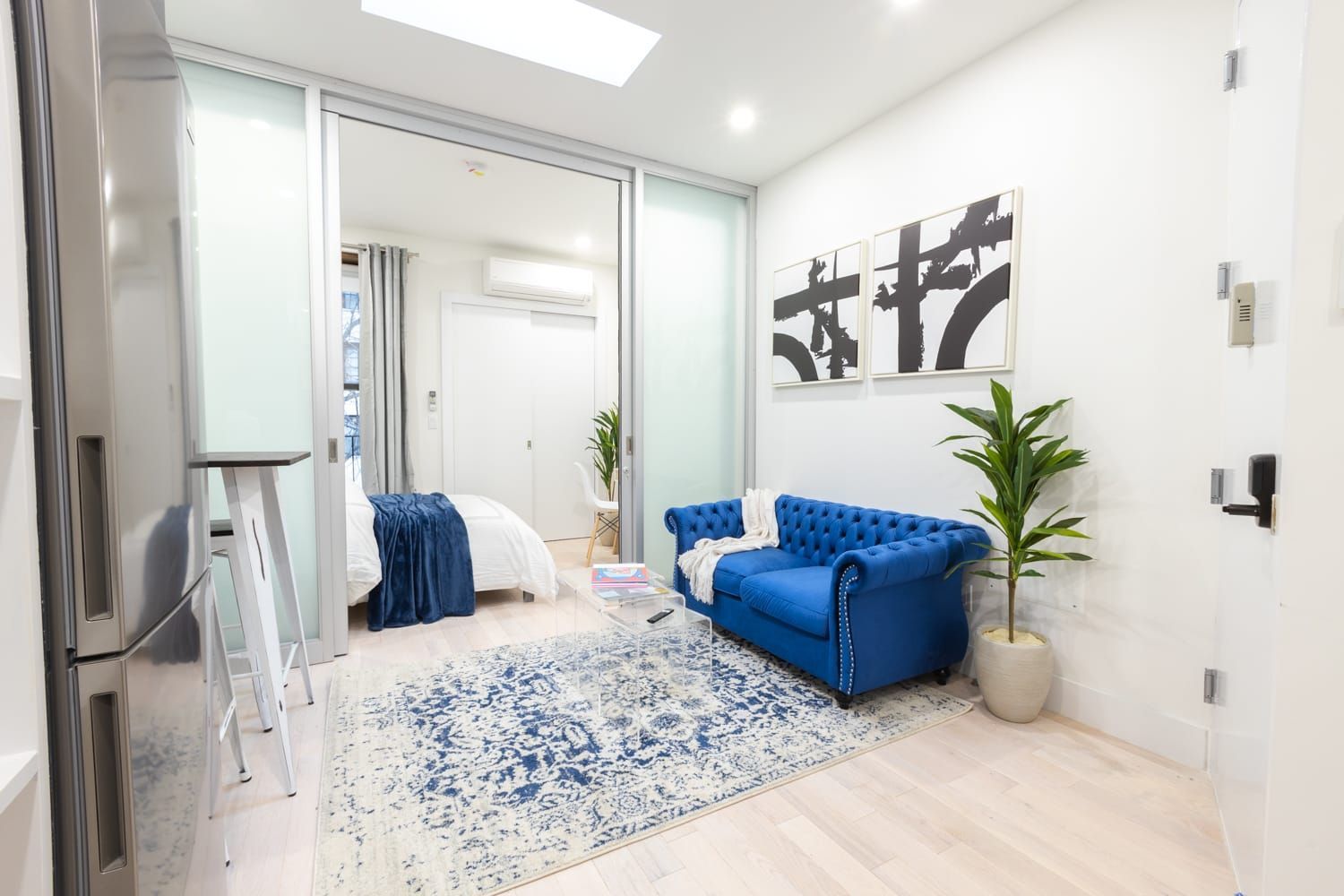 1-bedroom apartment with sofa, coffee table, white bed, wall art, frosted glass doors. The apartment has a dishwasher/dryer.