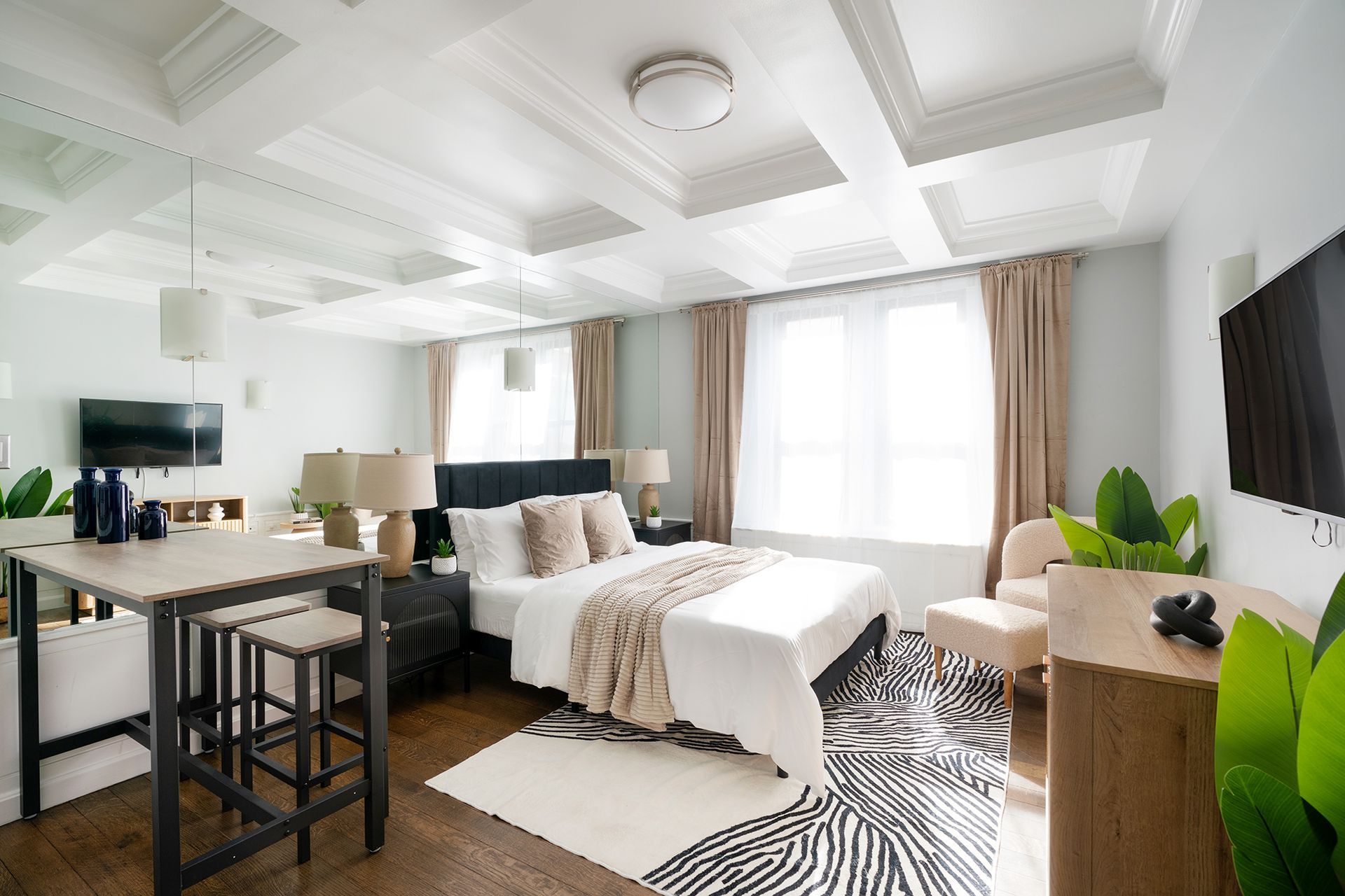 Contemporary studio apartment with a white bed, beige sofa, round wooden coffee table, potted plant, wall-mounted TV, and large windows with beige curtains. The apartment includes a doorman and gym and is located in the Upper West Side.