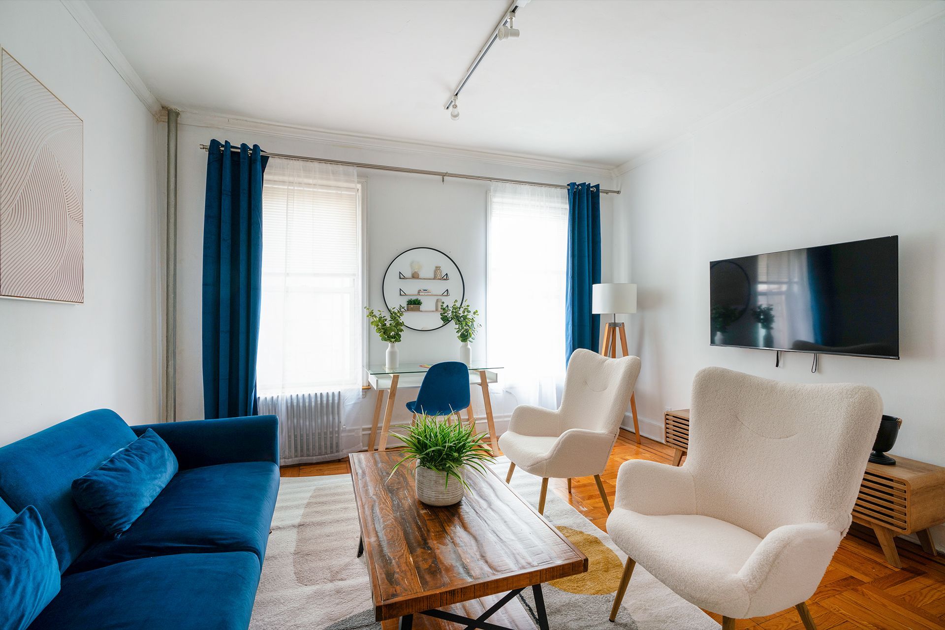 Bright one-bedroom apartment with a blue sofa, white armchairs, wooden coffee table, potted plants, and large windows with blue curtains. The apartment is located in Midtown East.