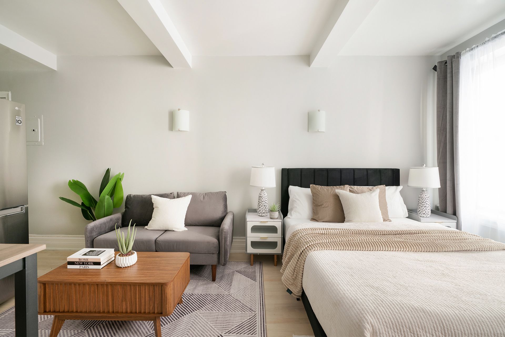 Bright studio apartment with a beige bed, beige sofa, round wooden coffee table, dining area with a table and chairs, potted plant, and large windows with beige curtains. The apartment includes a doorman and gym and is located in the Upper West Side.