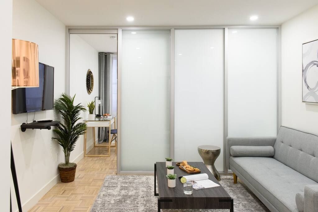 Bright one-bedroom apartment with a gray sofa, glass coffee table, TV mounted on the wall, potted plant, breakfast bar with stools, and large frosted glass doors. The apartment has a washer and dryer and an in-unit elevator.