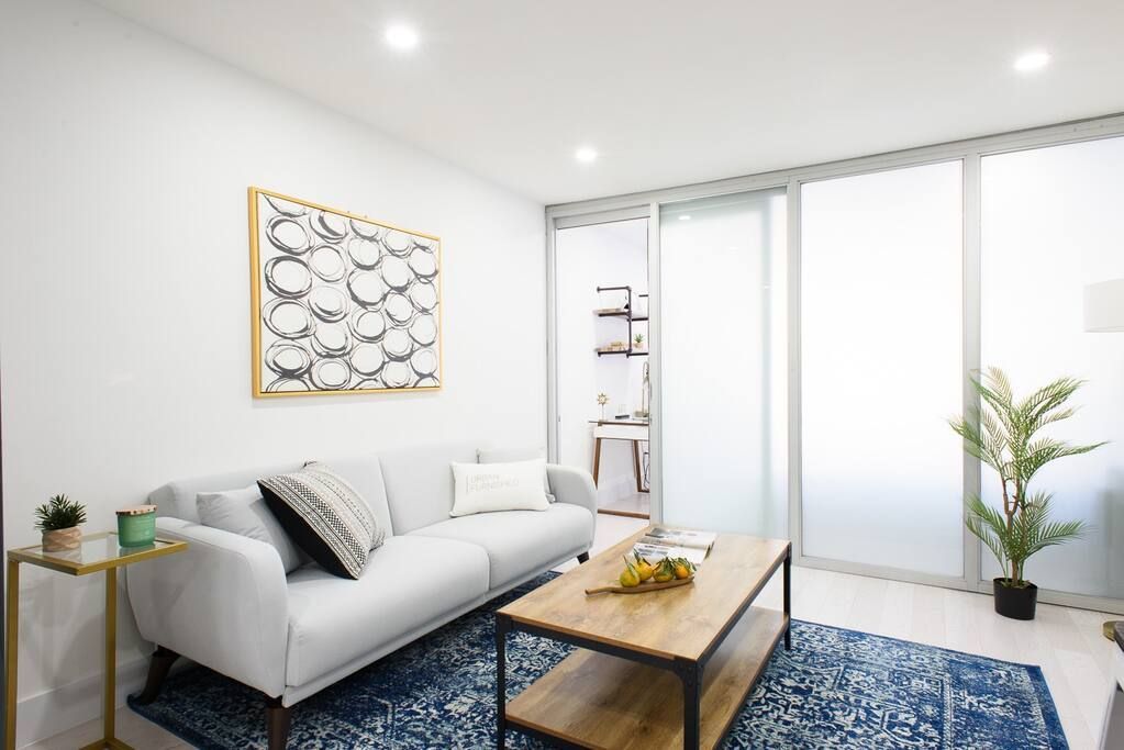 Modern one-bedroom apartment with a white sofa, wooden coffee table, blue patterned rug, potted plants, large frosted glass doors, and abstract wall art. The apartment has a washer and dryer and an in-unit elevator.