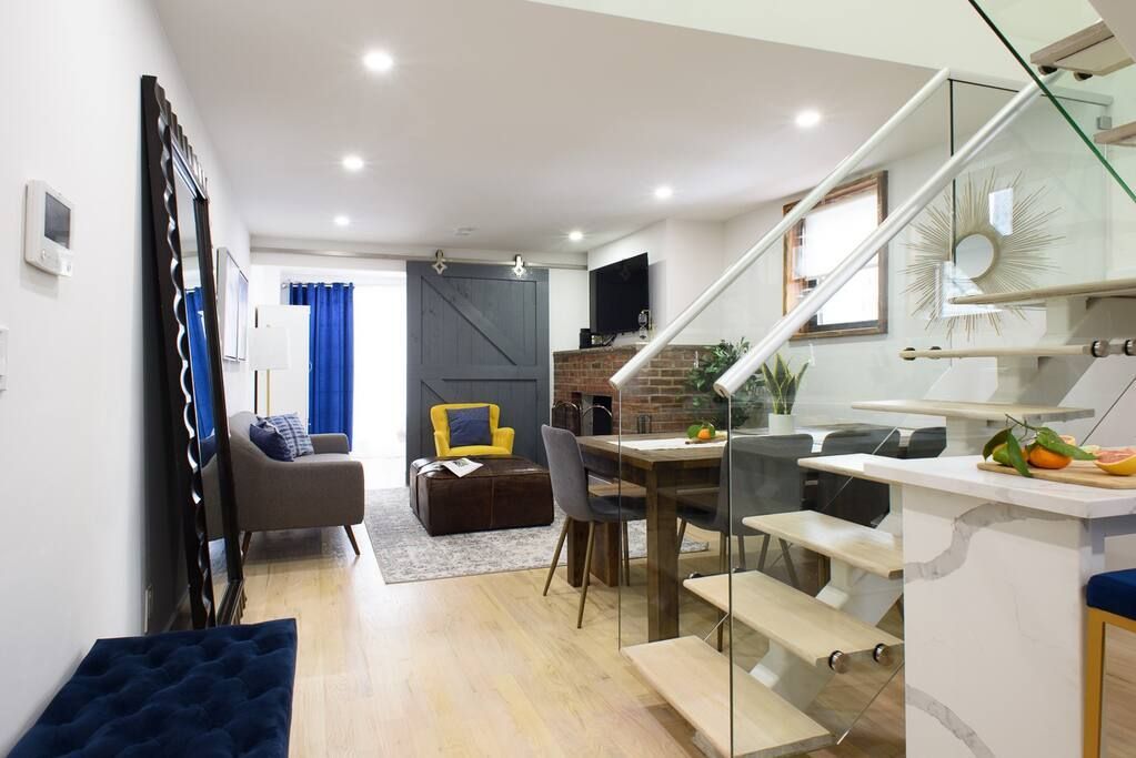 Modern three-bedroom apartment with a white kitchen island, glass staircase, black dining chairs, gray sofa, and yellow armchair. The apartment has a washer and dryer and an in-unit elevator.