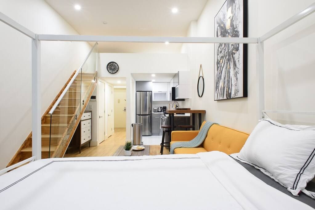 Modern two-bedroom apartment with a white bed, yellow sofa, abstract black and white wall art, kitchen area with stainless steel appliances, and stairs leading to a second level. The apartment has a washer and dryer.