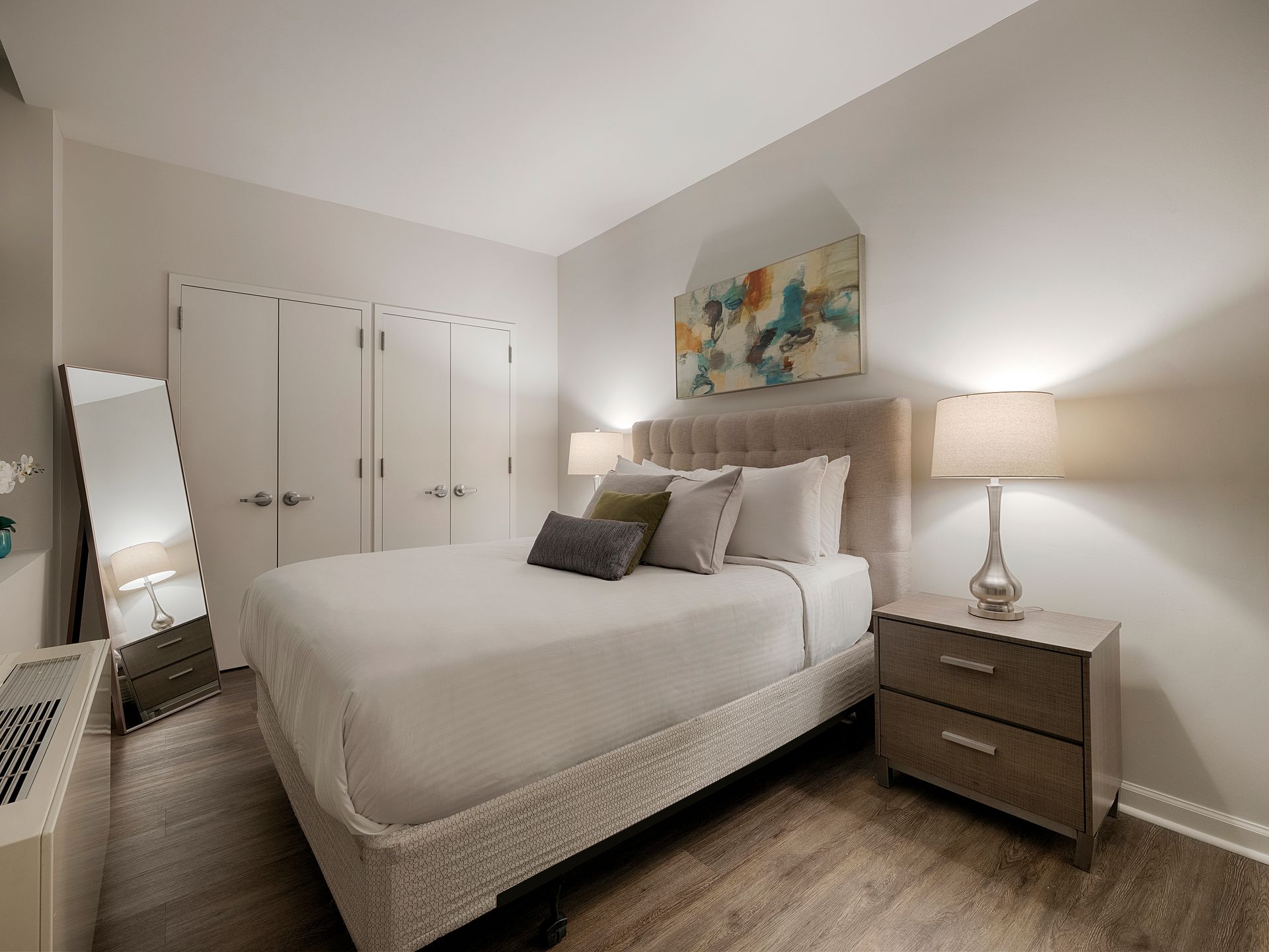 Cozy 1-bedroom apartment in the Financial District featuring a comfortable double bed with a cushioned headboard, neatly dressed in white linens. The room includes a bedside table with a lamp on each side, a wardrobe with double doors, and a serene piece of artwork above the bed. The space is warmly lit, creating a calm and inviting atmosphere.