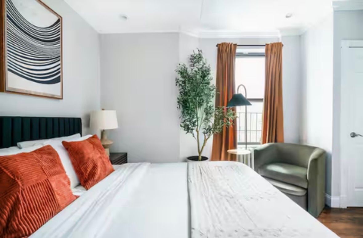 Bedroom with white walls, large bed, wood dresser, cloud mirror, and two windows with sheer curtains.