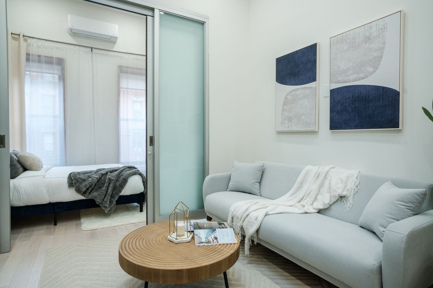 Contemporary one-bedroom apartment with a light gray sofa, round wooden coffee table, blue and gray wall art, and a frosted glass door. The bedroom has a white bed with a gray blanket. The apartment has a washer and dryer.