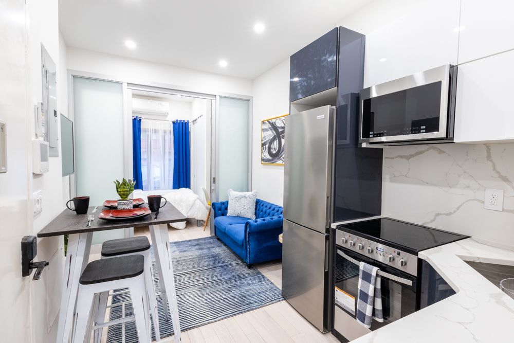 Modern one-bedroom apartment with a blue sofa, glass coffee table, white kitchen island with black bar stools, stainless steel appliances, and large windows with blue curtains. The apartment has a dishwasher and dryer.