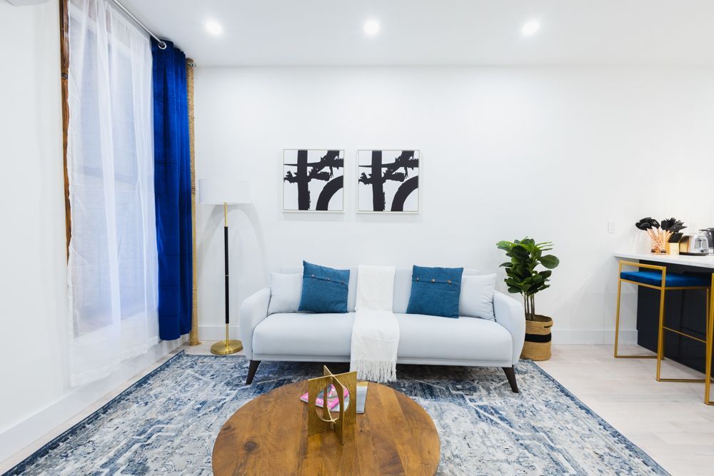 Contemporary one-bedroom apartment with a white sofa, blue patterned rug, wooden coffee table, abstract wall art, and large windows with blue curtains. The apartment has a washer and dryer.