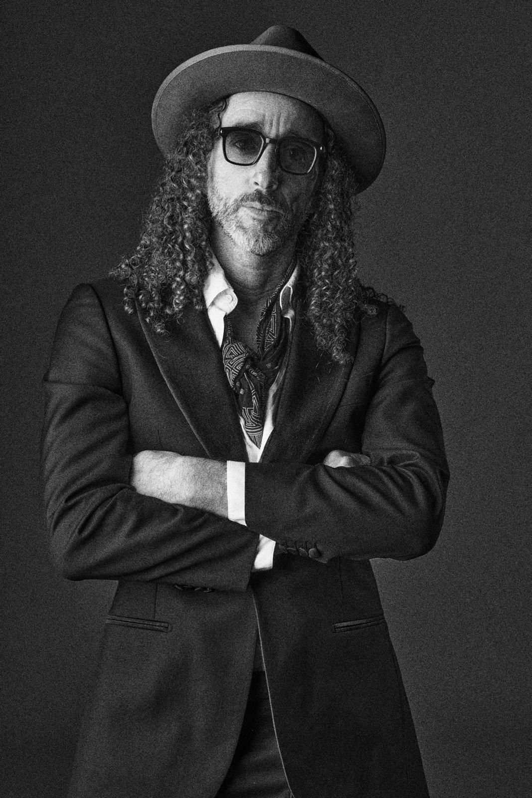 Man with long curly hair, wearing hat, suit, and scarf, arms crossed, looking at the camera.