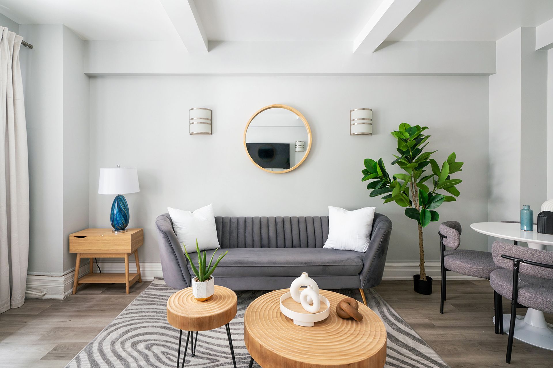Modern one-bedroom apartment with a gray sofa, round wooden coffee table, potted plant, wall-mounted TV, and large round wall mirror. The apartment includes a doorman and gym and is located in the Upper West Side.