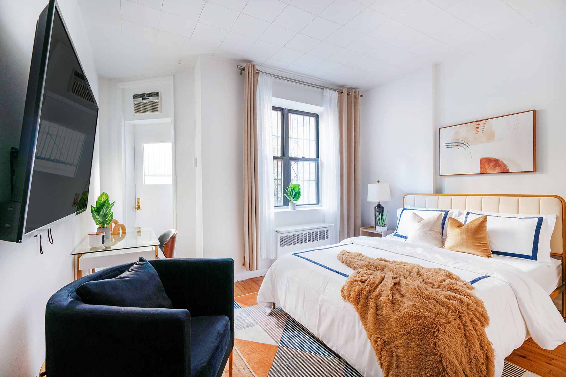 Modern studio apartment with a white bed, beige throw blanket, dark blue armchair, wooden desk with a chair, potted plant, and large windows with beige curtains. The apartment includes access to a gym and is located in the Upper West Side.