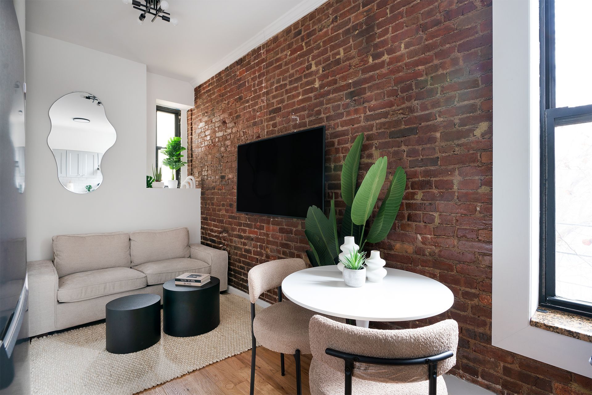 Newly renovated 2-bedroom apartment in the Lower East Side featuring an exposed brick wall, a cozy beige sofa, a round white dining table with two plush chairs, and a modern black coffee table set. The room is decorated with a large flat-screen TV and a stylish wall mirror.