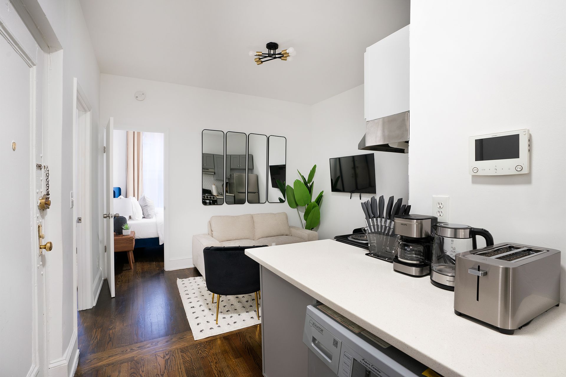 Modern three-bedroom apartment with a white kitchen, black chairs, potted plant, large wall mirror, and a cozy living area with a white sofa. The apartment includes a washer and dryer and is located in the Upper East Side.