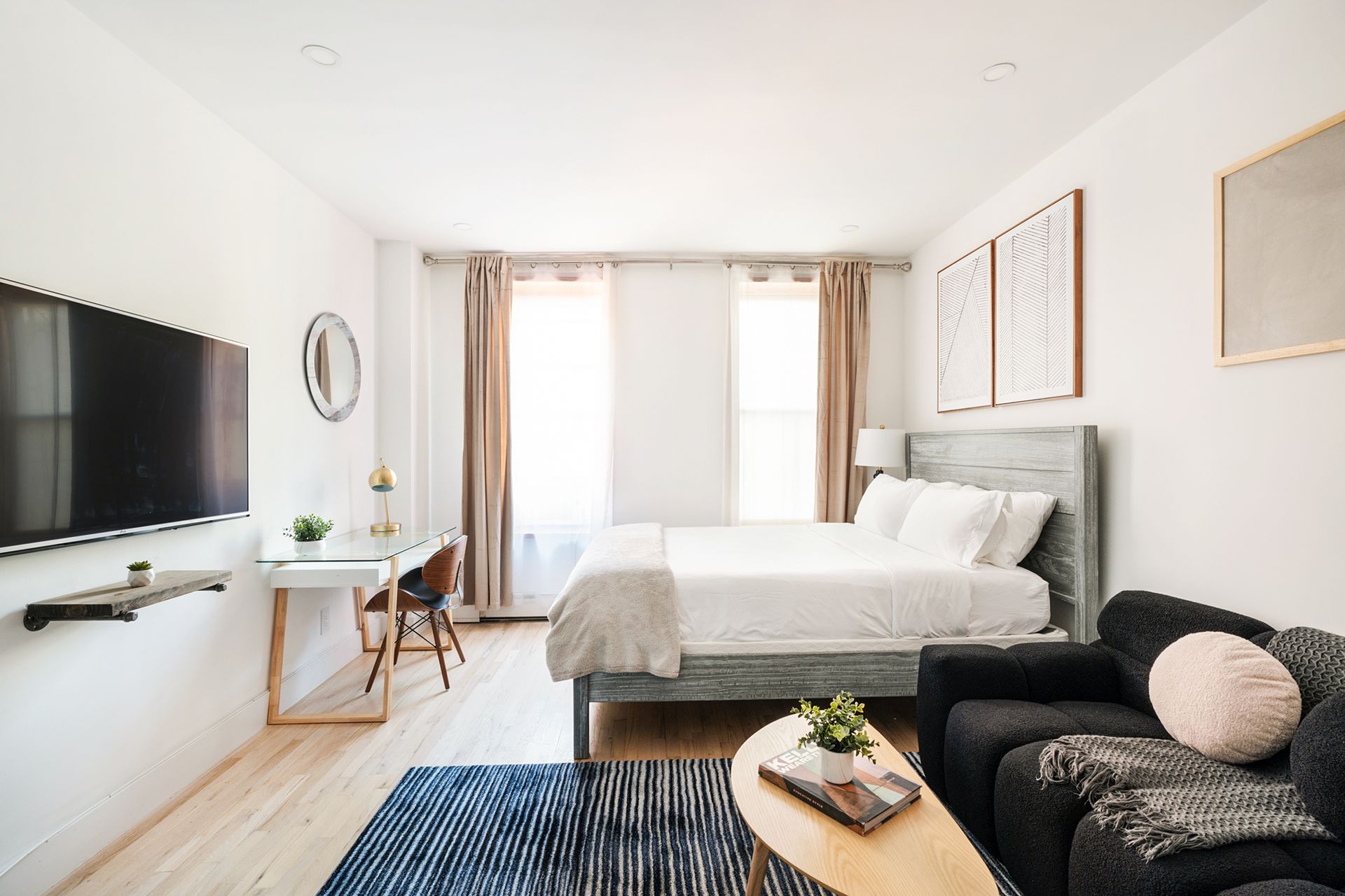 Newly renovated studio apartment with a white bed, yellow sofa, blue curtains, abstract black and white wall art, and a clear coffee table. The apartment has a dishwasher and dryer.