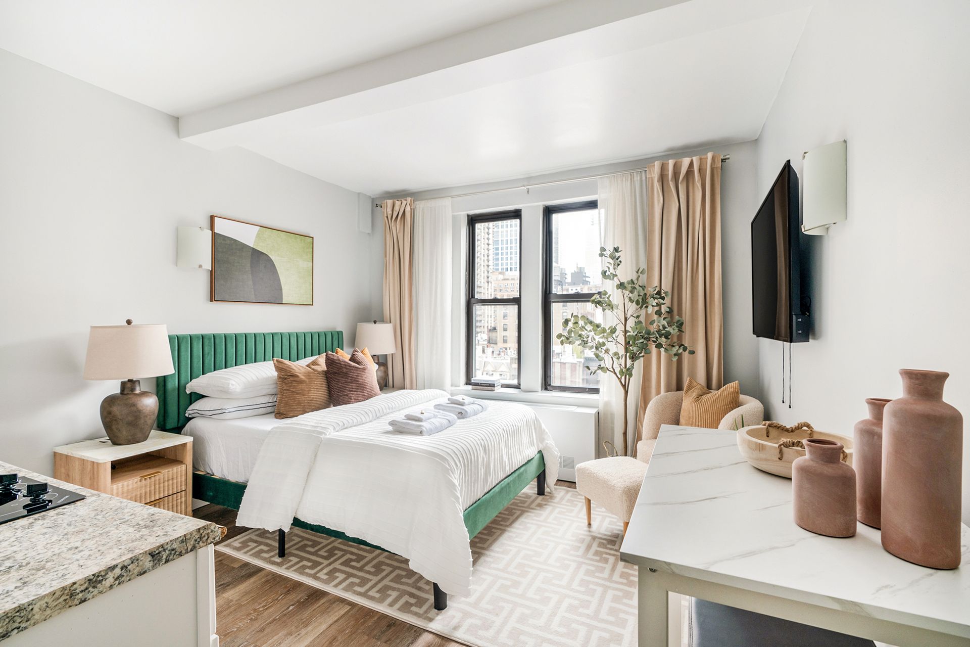 Spacious studio apartment with a beige bed, beige sofa, round wooden coffee table, dining area with a table and chairs, potted plant, and large windows with beige curtains. The apartment includes a doorman and gym and is located in the Upper West Side.