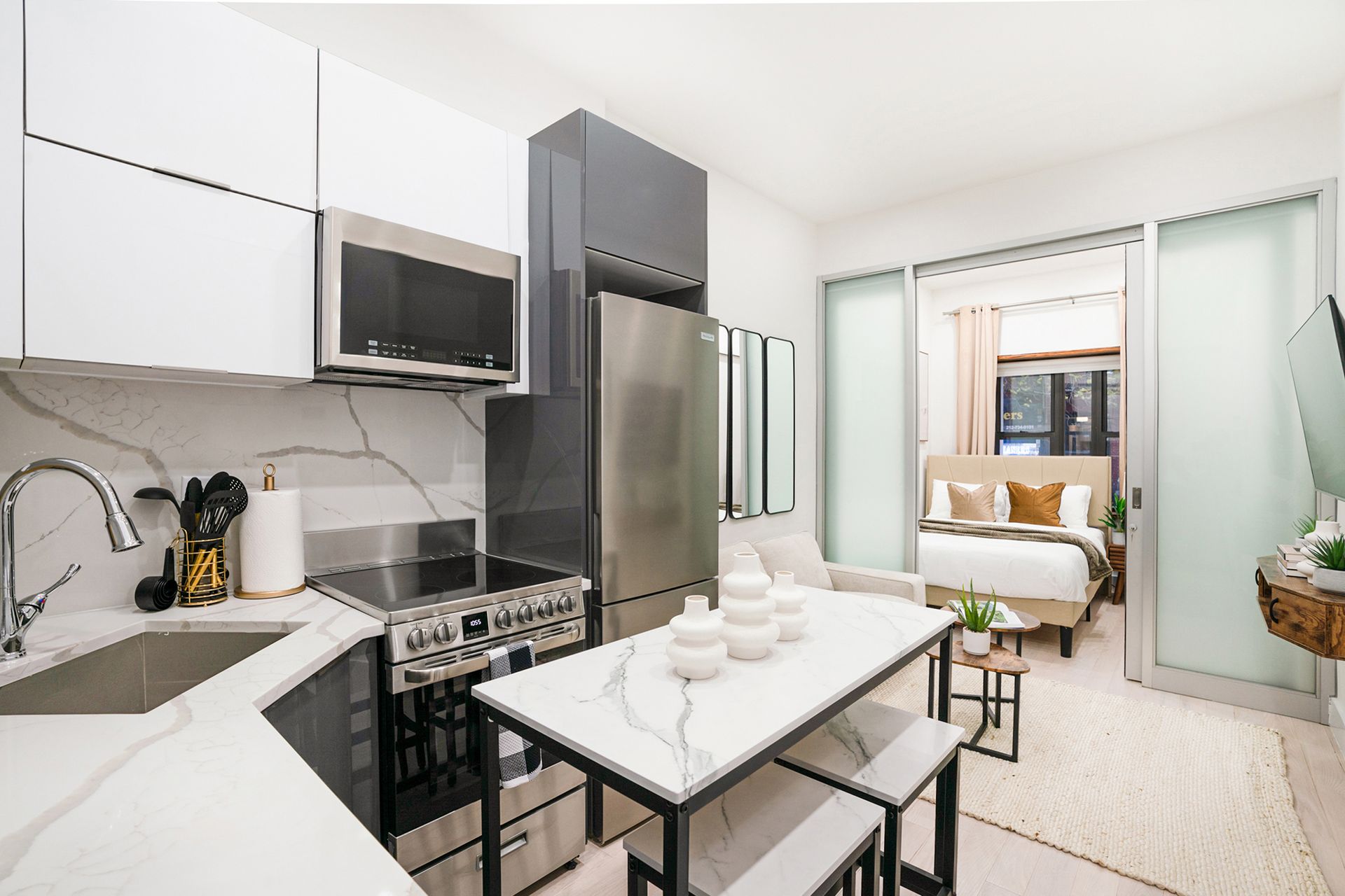 Contemporary one-bedroom apartment with a white bed, modern kitchen with stainless steel appliances, white countertops, and an open living area. The apartment has a washer and dryer.