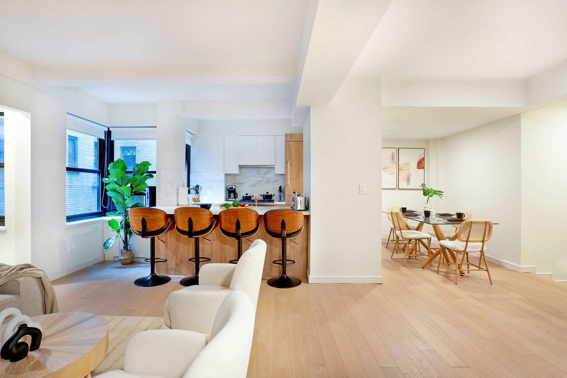 Spacious two-bedroom apartment with a modern kitchen featuring wooden cabinets, a breakfast bar with wooden barstools, potted plants, and large windows. The dining area has a round table with chairs and wall art. The apartment is located in Midtown East.