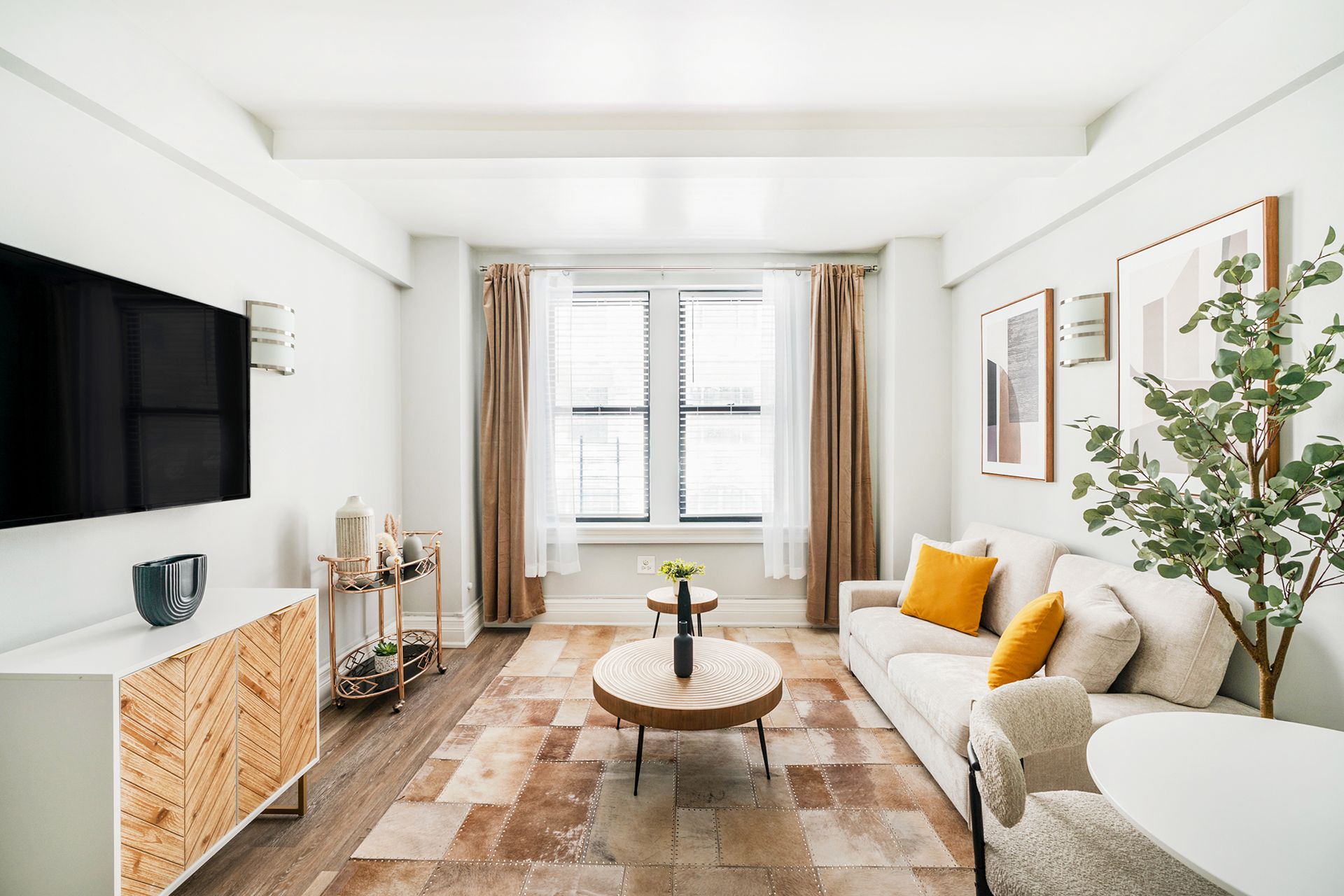 Modern one-bedroom apartment with a beige sofa, round wooden coffee table, wall-mounted TV, potted plant, and large windows with beige curtains. The apartment includes a doorman and gym and is located in the Upper West Side.