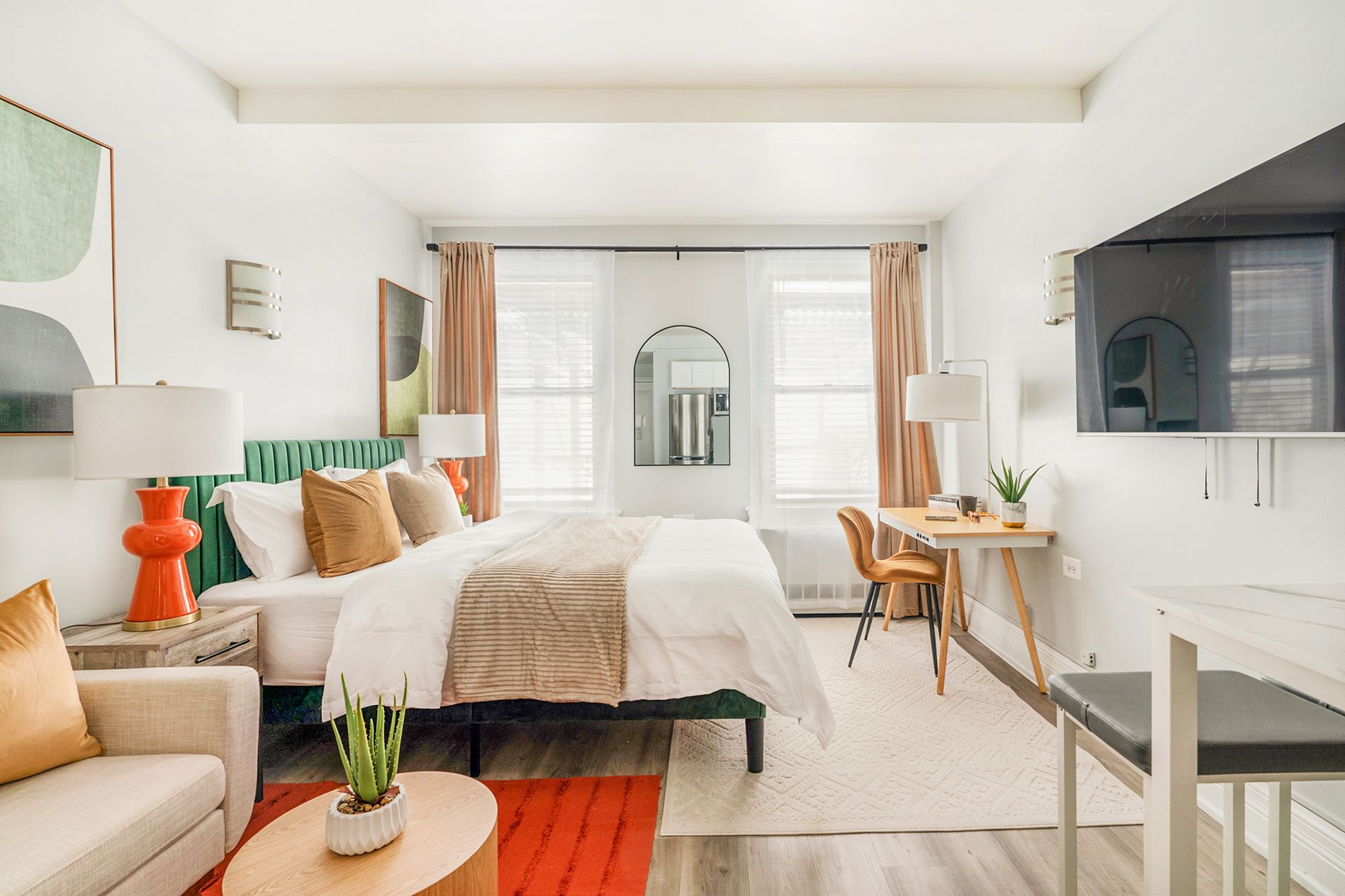 Bright Studio apartment with a white bed, bedside tables with lamps, wooden desk with a chair, potted plant, wall-mounted TV, and large windows with beige curtains. The apartment includes a doorman and is located in the Upper West Side.