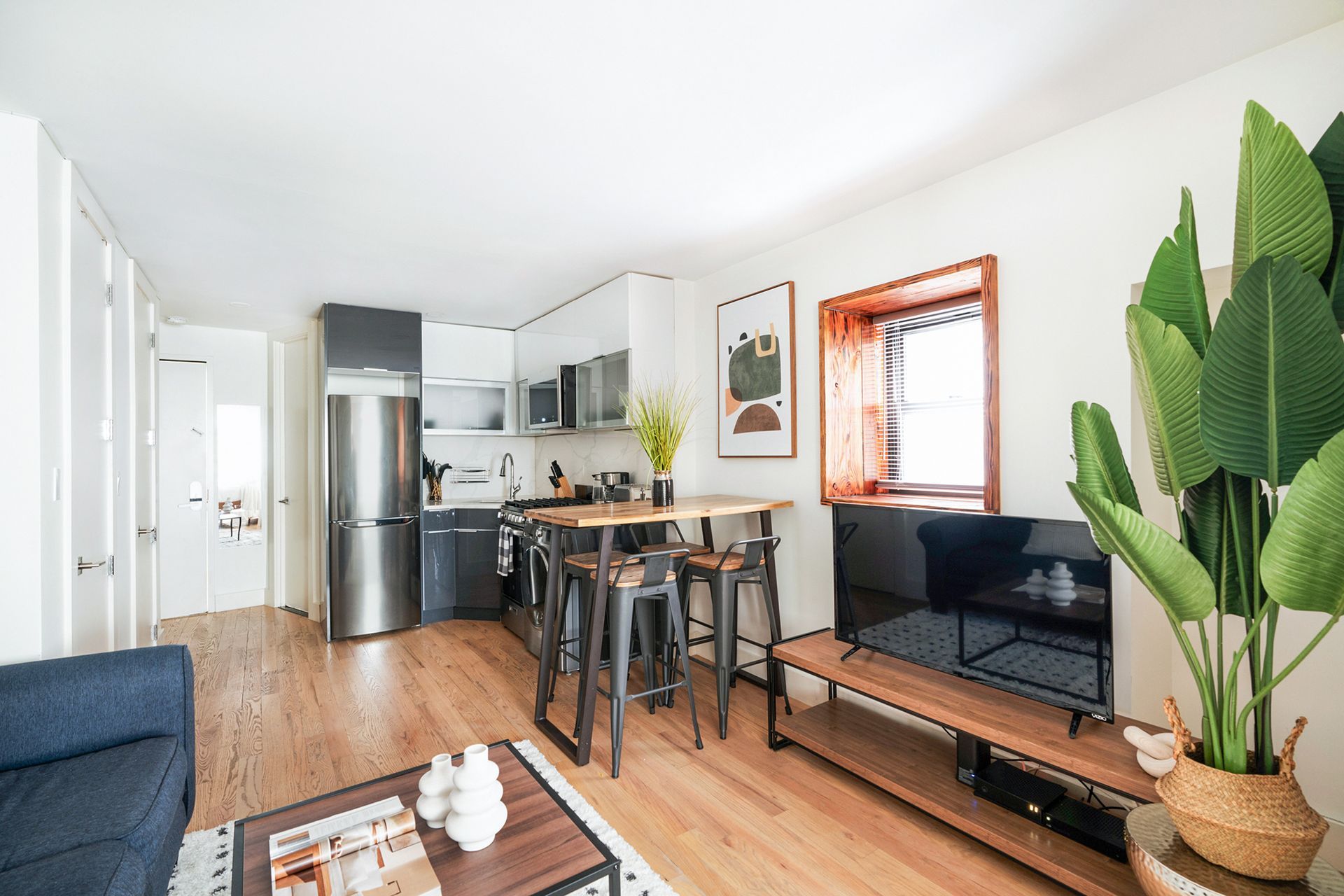Contemporary one-bedroom apartment with a white bed, blue and white patterned rug, kitchen area with a breakfast bar, dining table with chairs, potted plants, and wall art. The apartment has a washer and dryer and an in-unit elevator.