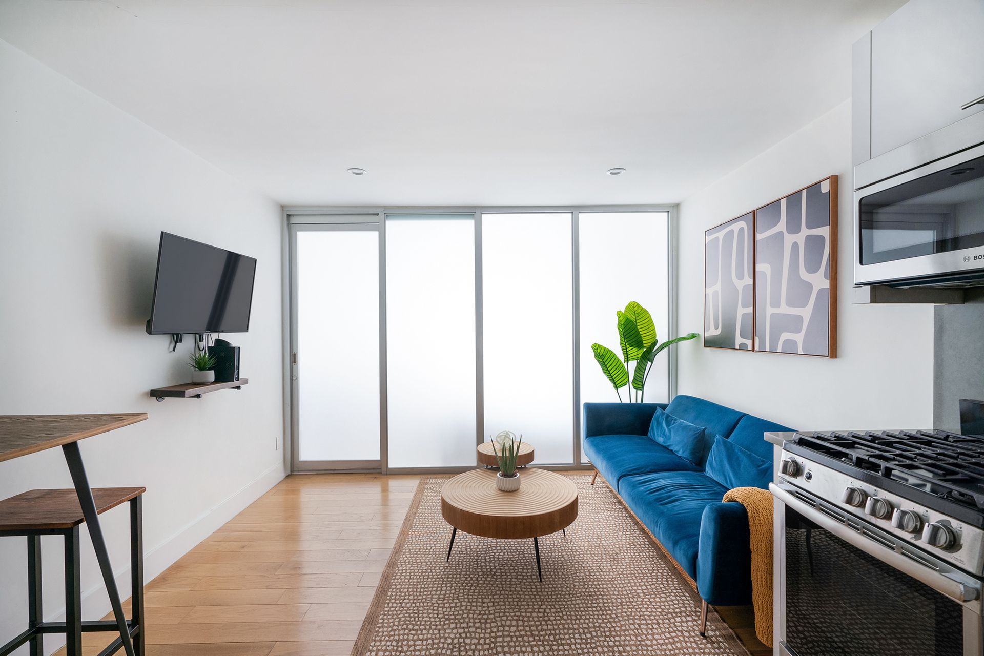 Modern one-bedroom apartment with a gray sofa, wooden coffee table, kitchen area with stainless steel appliances, stacked washer and dryer, and a breakfast bar with stools. The apartment has an in-unit elevator.
