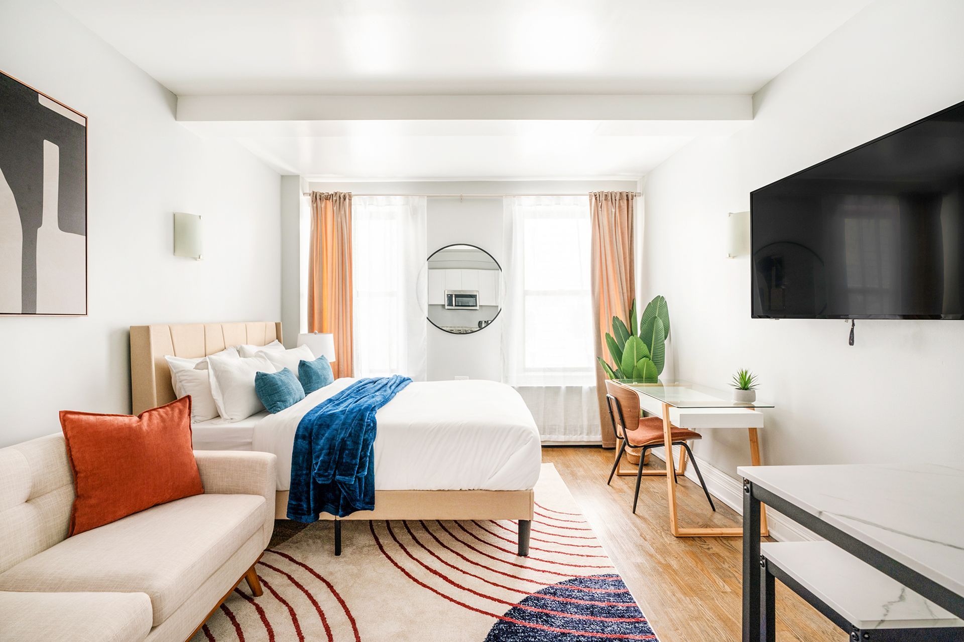 Modern studio apartment with a white bed, beige sofa, round wooden side table, potted plant, wall-mounted TV, and large windows with beige curtains. The apartment includes a doorman and gym and is located in the Upper West Side.