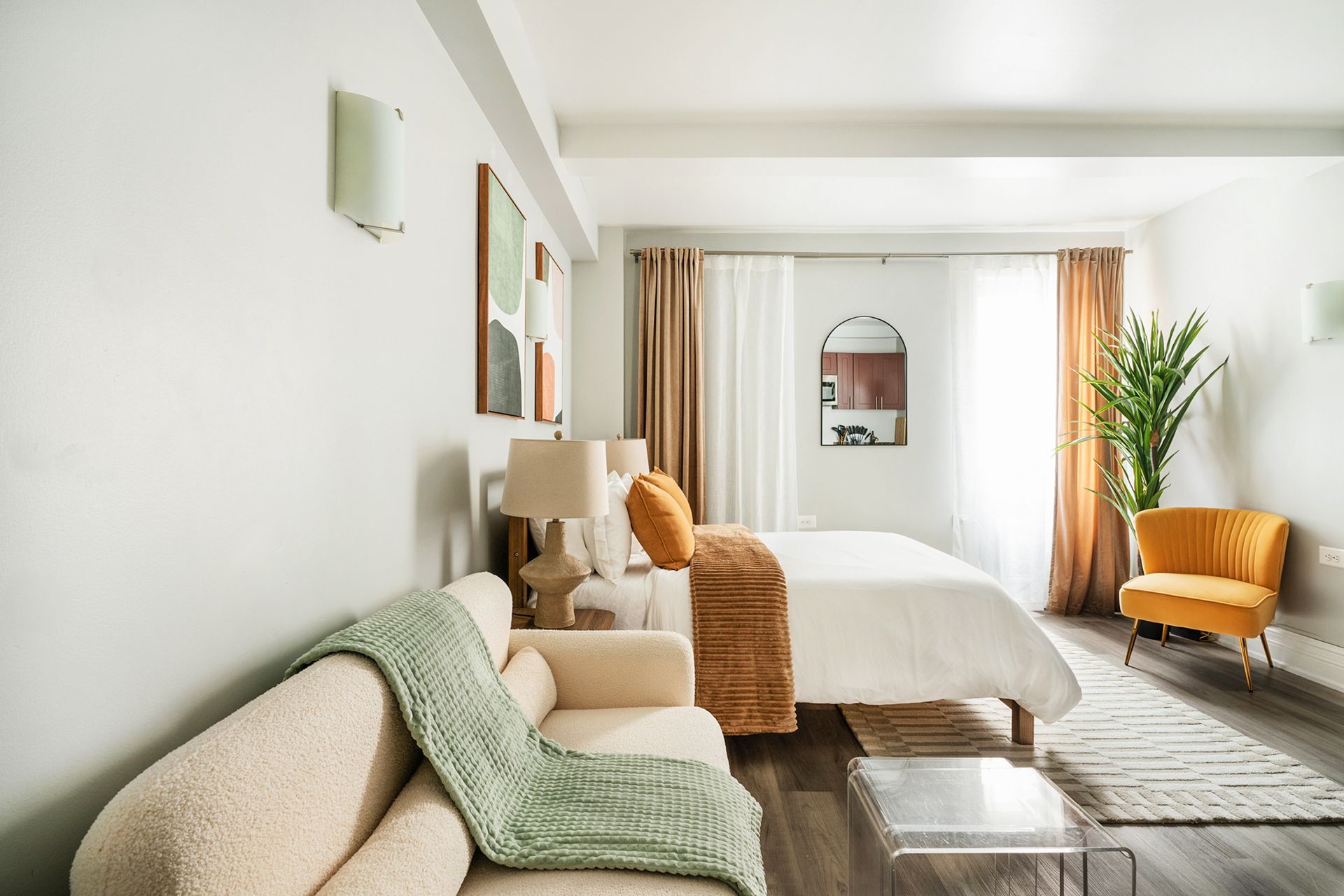 Modern studio apartment with a beige bed, beige sofa, round wooden coffee table, bedside table with lamp, potted plant, and large windows with sheer curtains. The apartment includes a doorman and gym and is located in the Upper West Side.