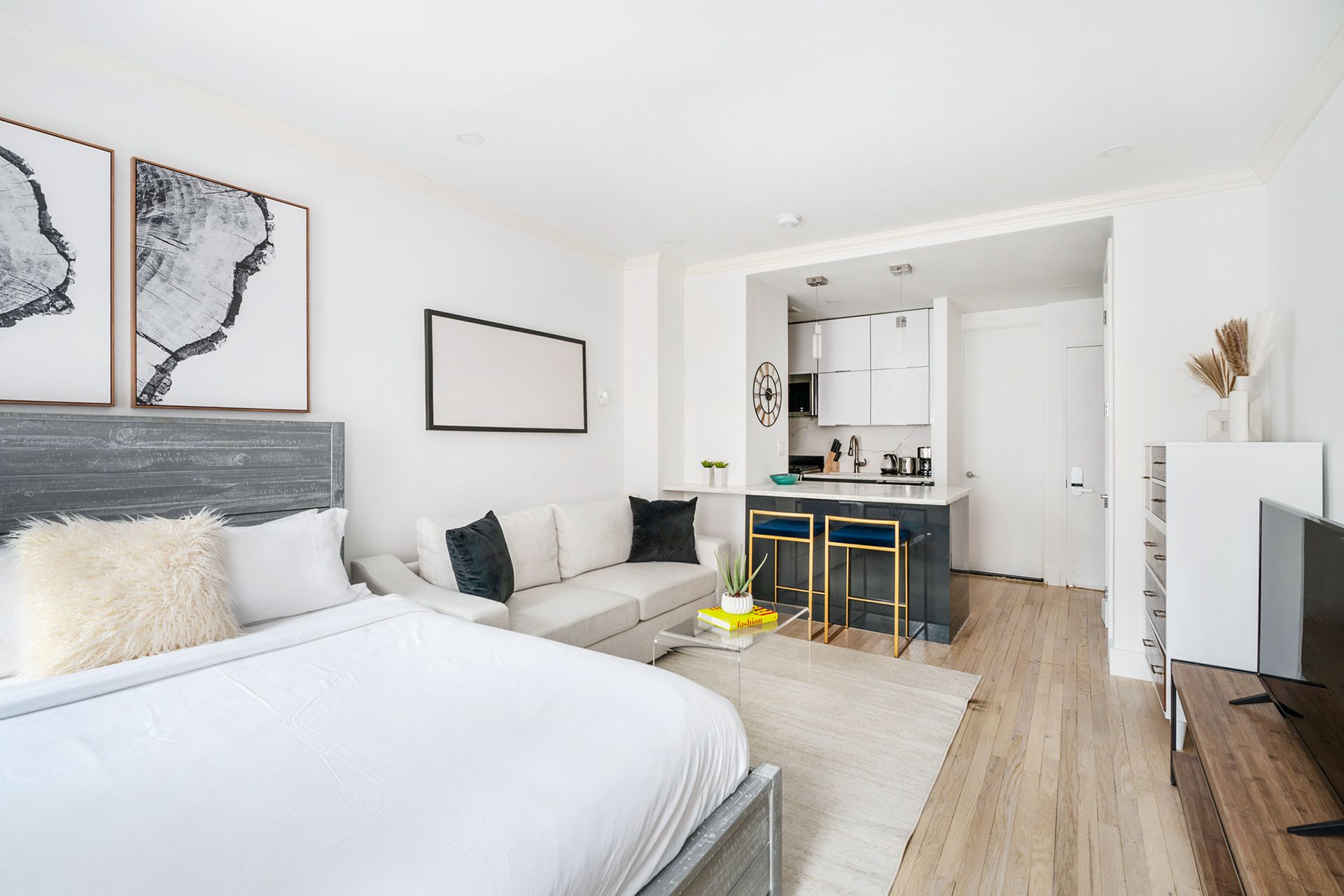 Bright studio apartment with a white bed, yellow sofa, gray patterned rug, large windows with white curtains, a small desk with a chair, and abstract wall art. The apartment has a washer and dryer.