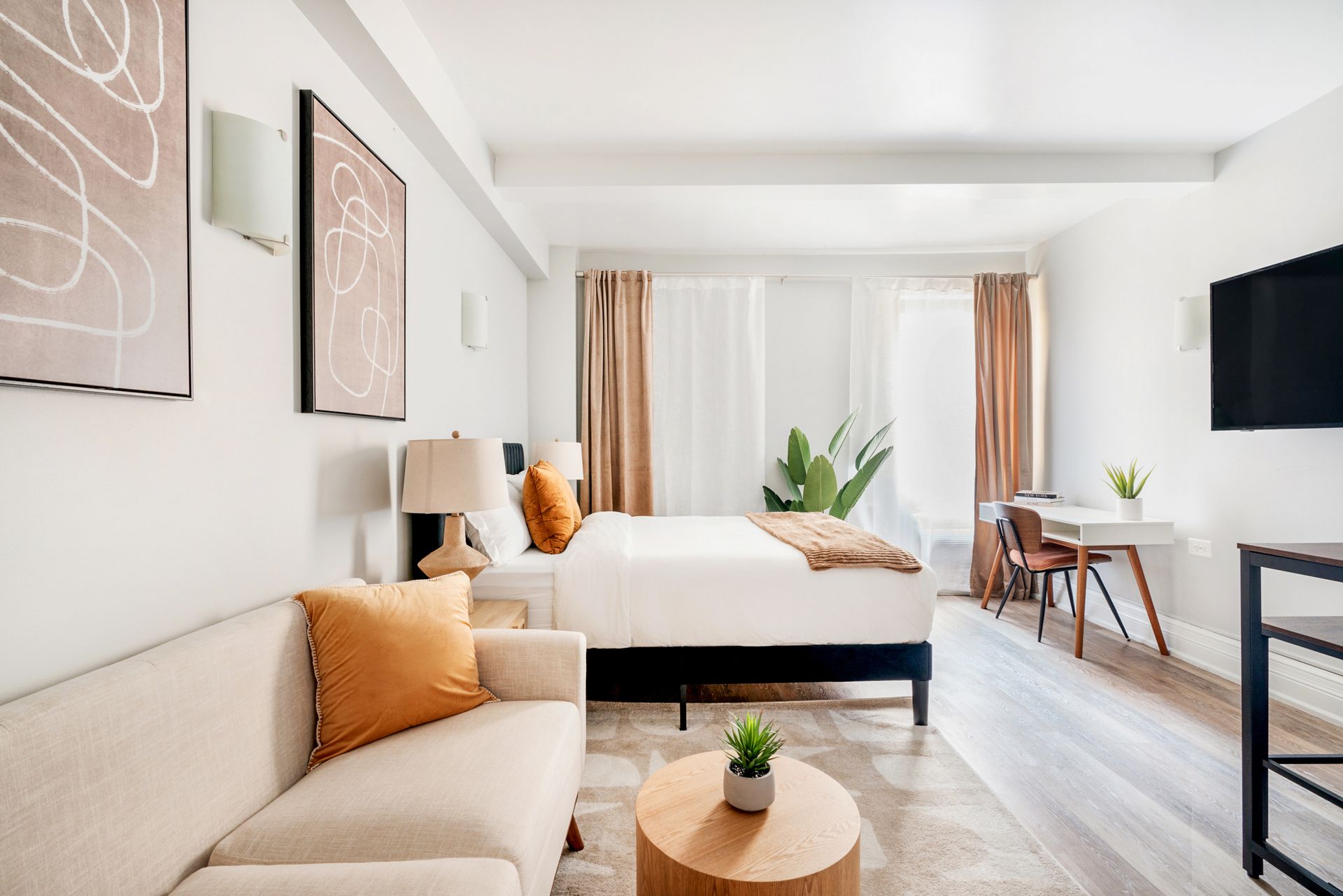 Bright studio apartment with a beige sofa, white bed, round wooden side table, potted plant, wall-mounted TV, and large windows with beige curtains. The apartment includes a doorman and gym and is located in the Upper West Side.