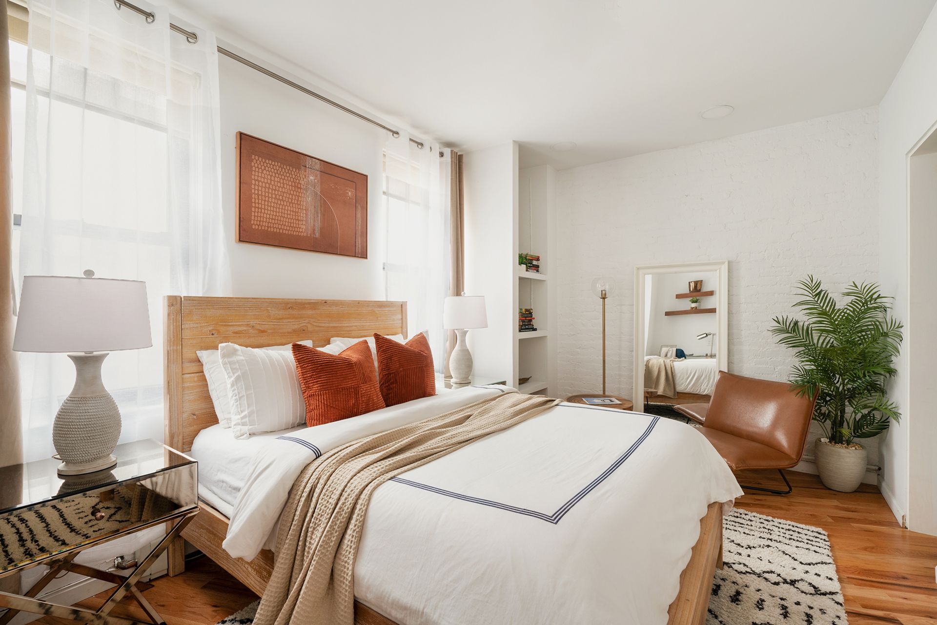 Charming 2-bedroom apartment in the West Village with a cozy and inviting bedroom. The room features a wooden bed frame with white and orange bedding, a tall plant, and a modern bedside table with a lamp. The space is adorned with warm-toned artwork, a leather chair, and a full-length mirror, creating a comfortable and stylish environment.
